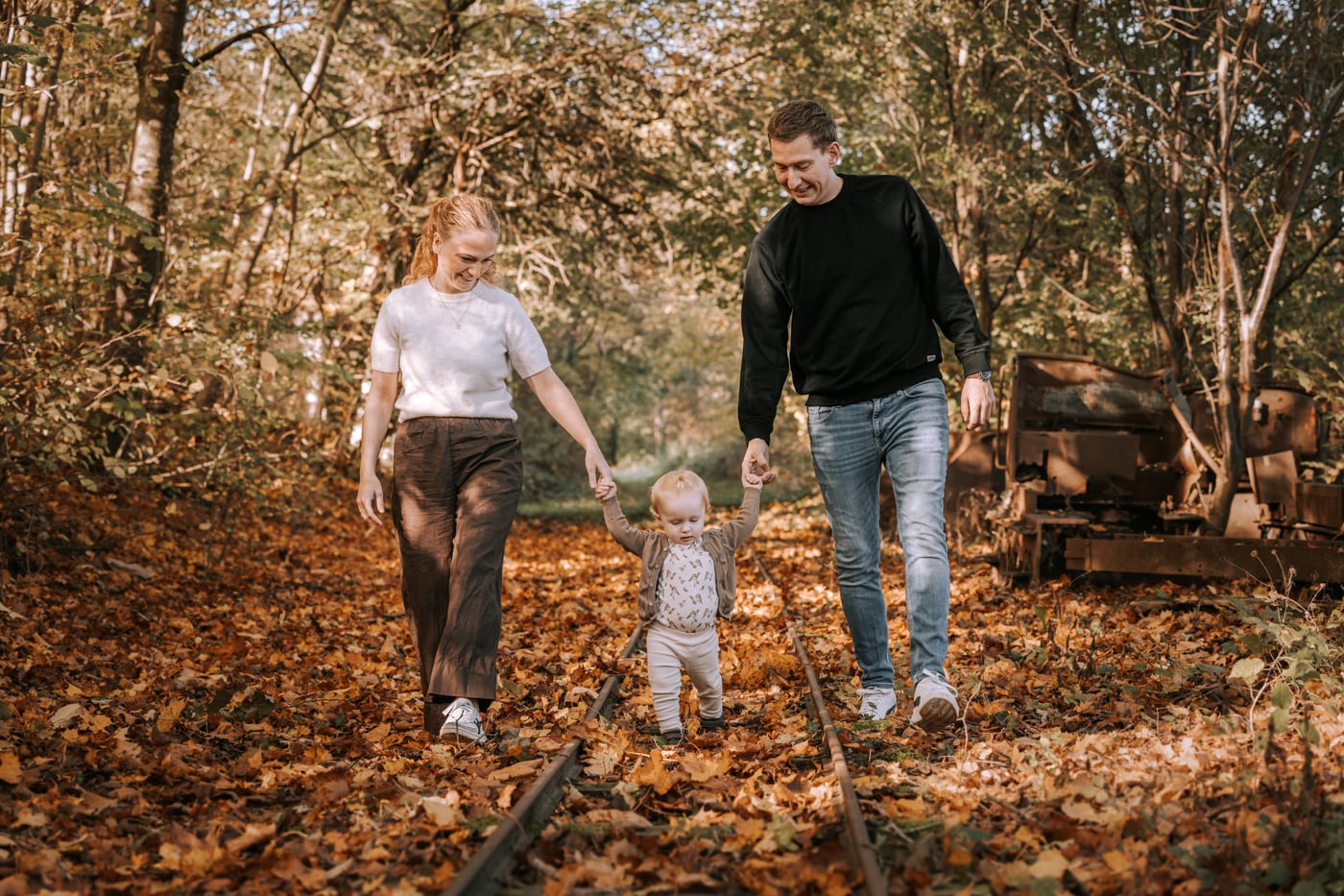 Mor og far holder deres lille barn i hænderne på en gåtur ad gamle skinner dækket af orange efterårsblade. Autentisk familiefotografering i Jylland.